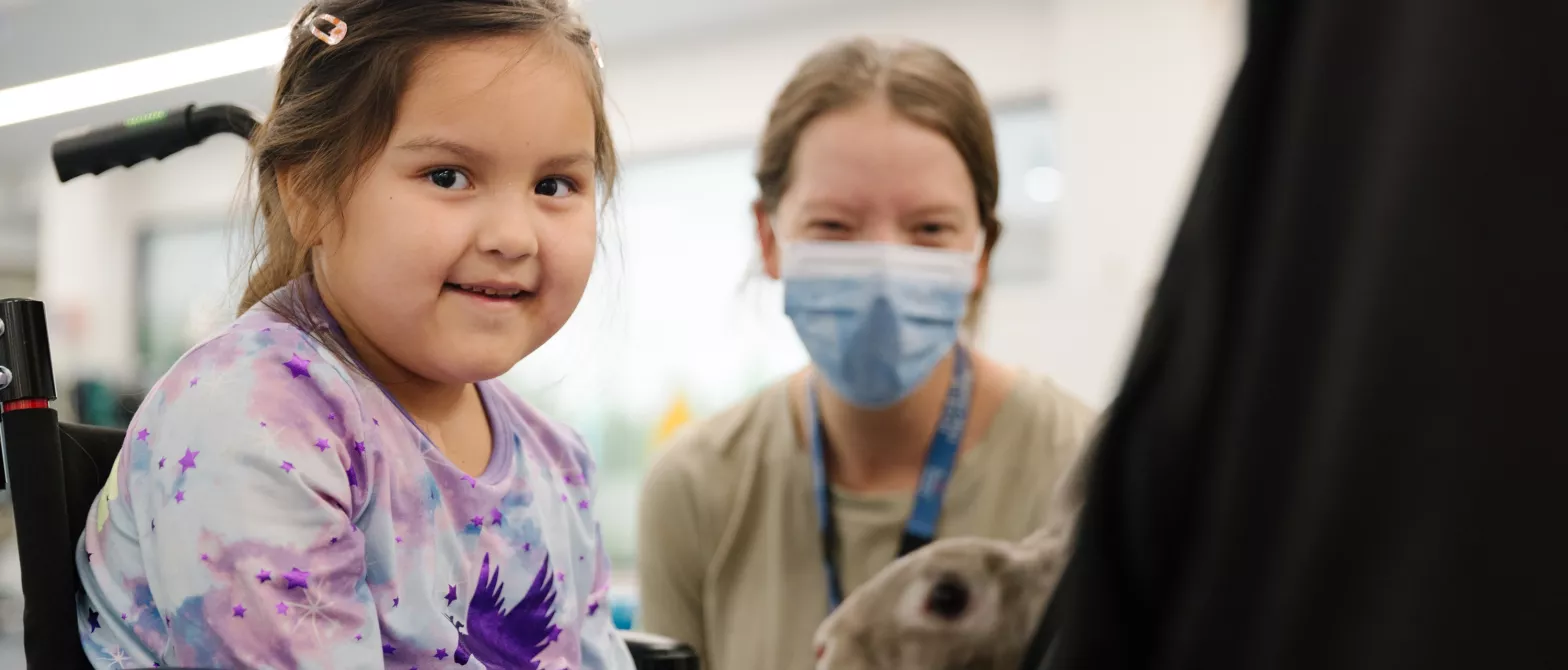 Child in wheelchair petting a rabbit, with staff member in background