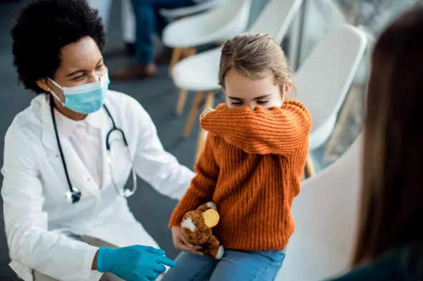 Child sneezing into elbow, being comforted by a doctor wearing a mask